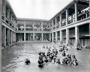 Vandret, sort / hvidt fotografi af en swimmingpool med to etagers balkon understøttet af søjler. Tilskuere ser svømmerne fra balkonen på andet niveau